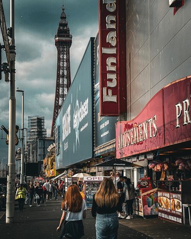 A busy scene showing people walking along the Promenade, with Funland in the foreground and the Blackpool Tower in the background