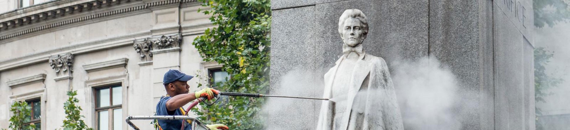 A specialist contractor standing on a cherry picker and cleaning a grey stone memorial to Edith Cavell using a jet wash.