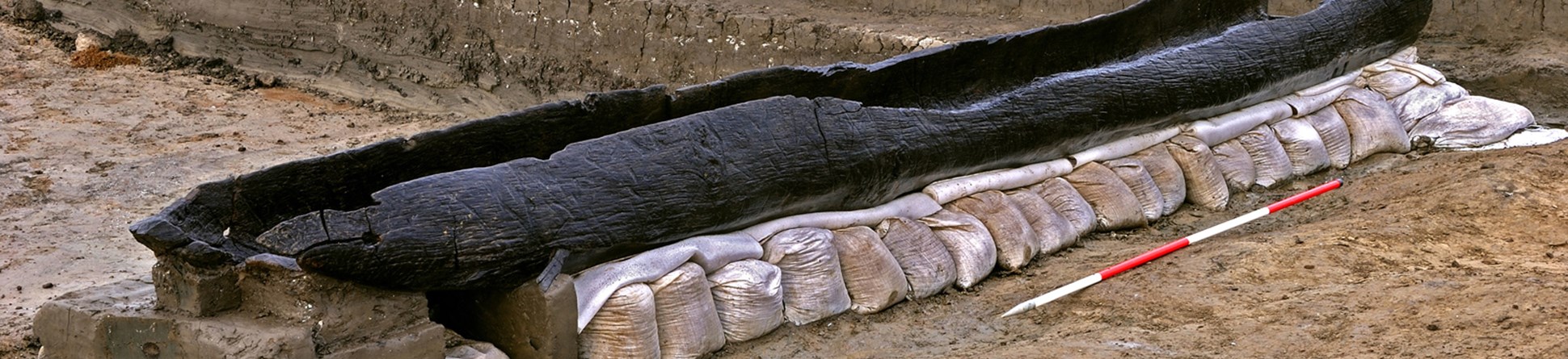 A hollowed, blackened tree log resting on a line of sand bags with a measuring stick alongside.