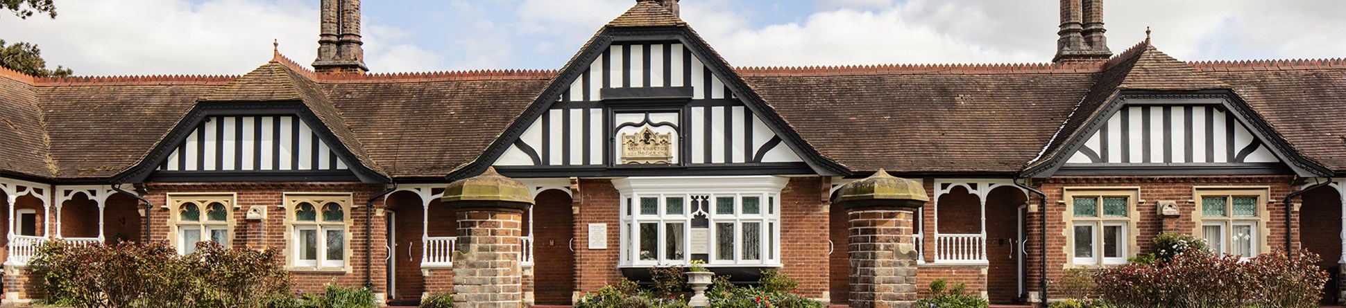 A low-level brick building with three gables with half-timbered decoration in black and white and pairs of star-topped chimney stacks.