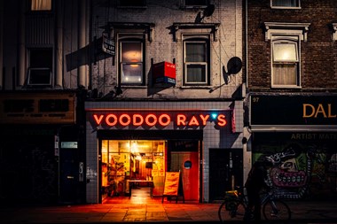 A photograph of a restaurant at night. The interior is lit yellow and the sign above reads 'Voodoo Ray's' in red light. 
