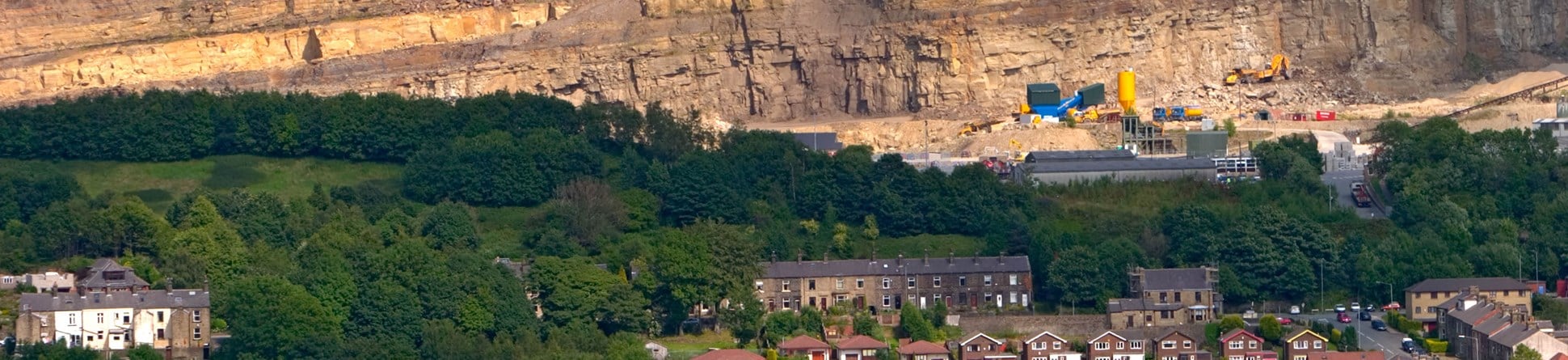A wide view of a broad quarry face, quarry machinery, woodland and rows of houses.  