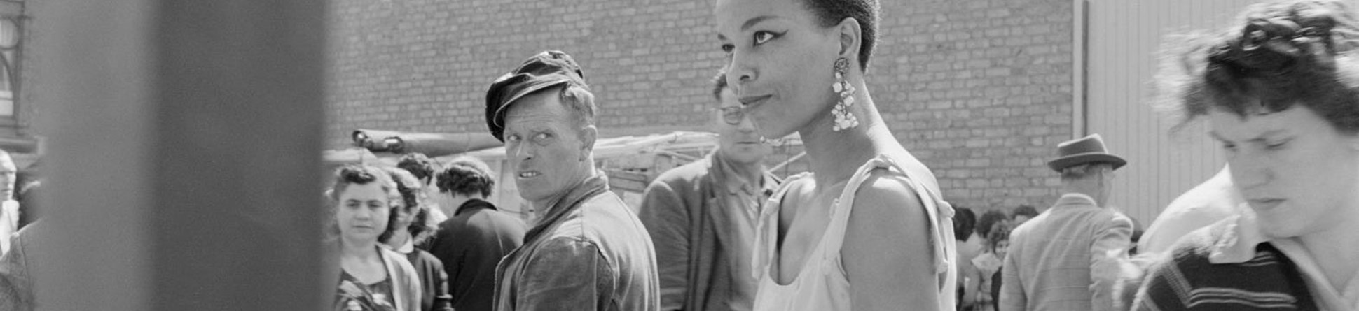 An informal portrait of a young black woman buying fruit at an unidentified open air market in north london, with a man looking back over his shoulder at her.