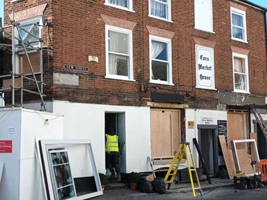 A view across a street to a three-storye red-brick building with white sash windows. The ground floor of the building is behind hoardings as it is being repaired.