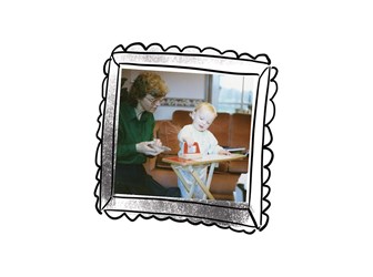 Photo of child playing with toy iron and ironing board displayed inside a hand-drawn frame. The child's mother kneels to the left, watching her. An older child stands on the right, only visible from knees up to chin.