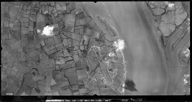 A black and white vertical aerial photograph featuring an airfield with three runways, set amongst a patchwork of fields, and sited adjacent to a wide river.