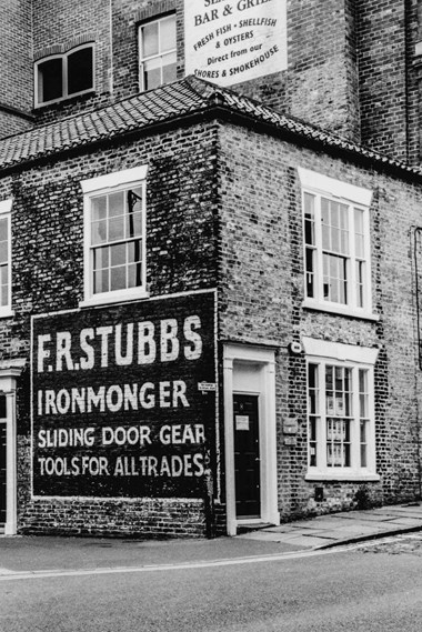A photograph of a street corner. On one side of the wall, a large painted advertisement reads, “F.R. STUBBS IRONMONGER SLIDING DOOR GEAR TOOLS FOR ALL TRADES”. On the other side of the wall, there is an entrance to a business. 