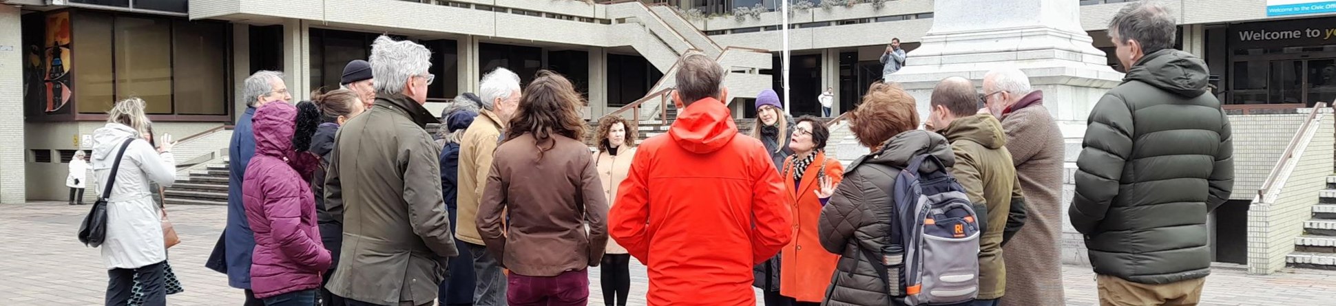 A group of people in warm coats stand listening to a woman speaking.