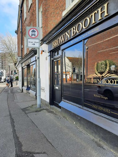 A side view of the oustide of a historic shopfront with full height windows. The woodwork and fascia are painted black with gold lettering.