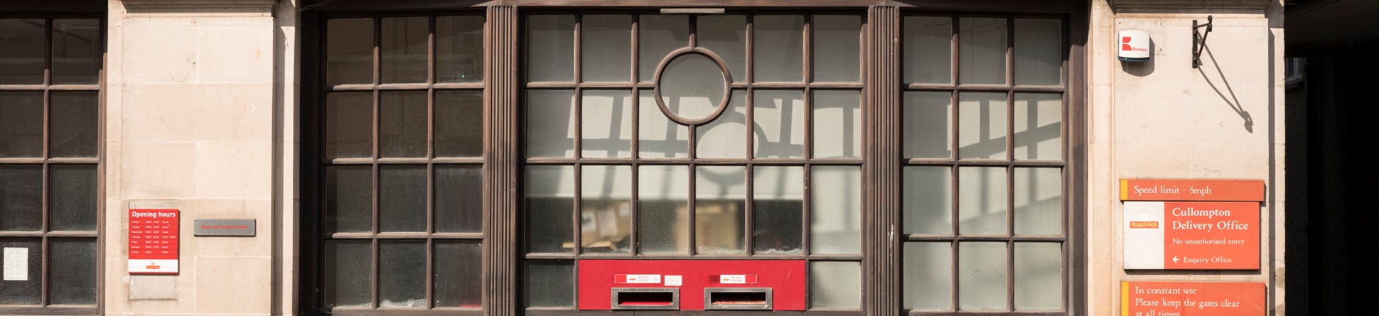 The front of a post office building with a large, multi-paned window above post-boxes which have been set into the masonry.