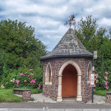 A circular stone bus stop with a white stone archway entrance, a tiled pointed roof, and a weather vane.