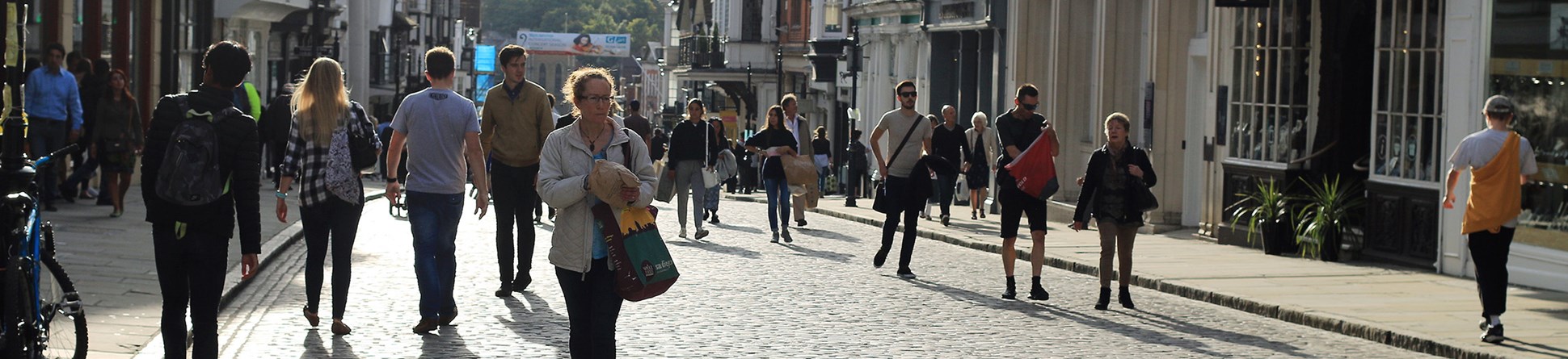 Guildford high street on a busy and sunny day. Pedestrians are walking both on the pavements and down the middle of the cobbled street.