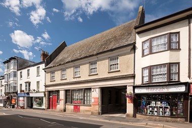 A view across a high street to a two story stone building. It has sash windows on the first floor and an elaborate window and red, panelled front door to the ground floor. There is also an archway allowing vehicles to deliver to the sorting office behind. It is a historic post office building.