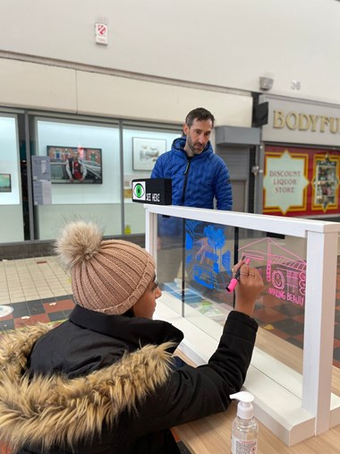 Girl wearing a bobble hat and anorak drawing on a glass pane watched on by a man