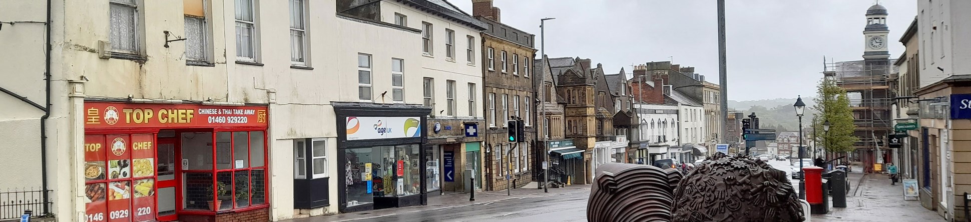 A historic high street on a wet day. In the foreground is a block of possibly sandstone and two large artworks of rusted metal.