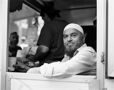 A black and white portrait photograph of a man sitting inside his food van. A person can be seen serving someone at the counter behind him. 